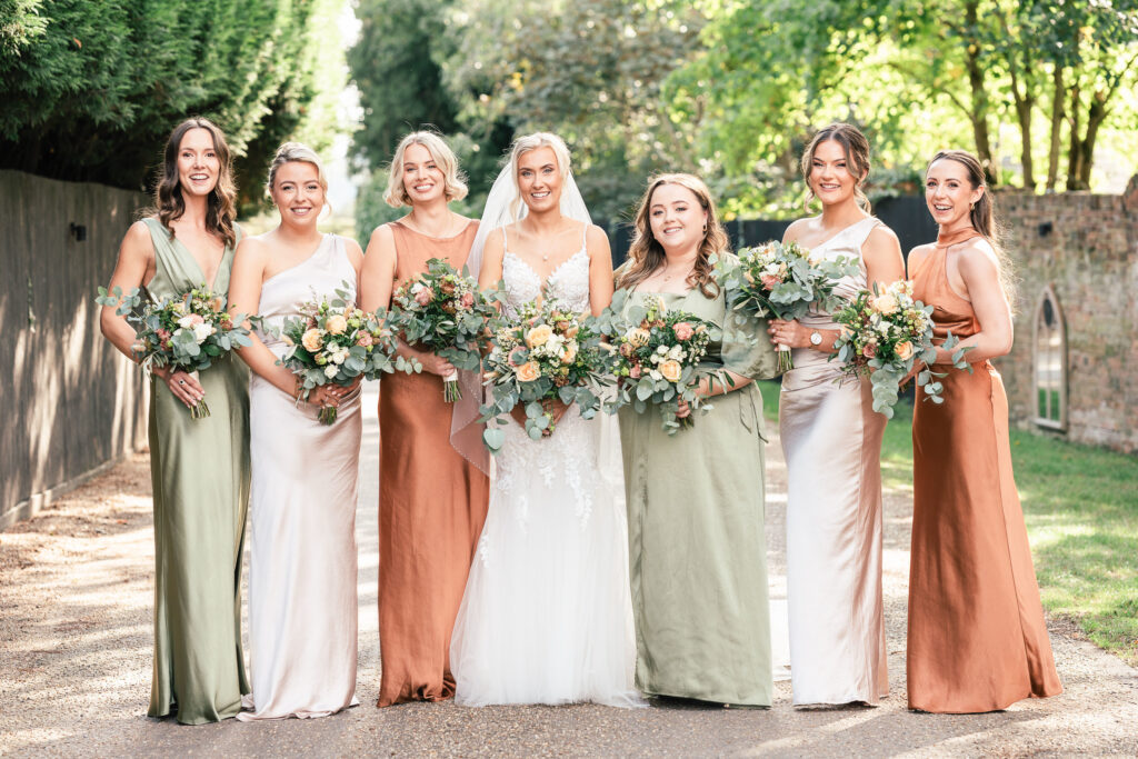 A bride in a white gown stands outdoors with six bridesmaids in green, ivory, and rust dresses. They are all smiling, holding bouquets of flowers, and surrounded by trees on a sunny day.
