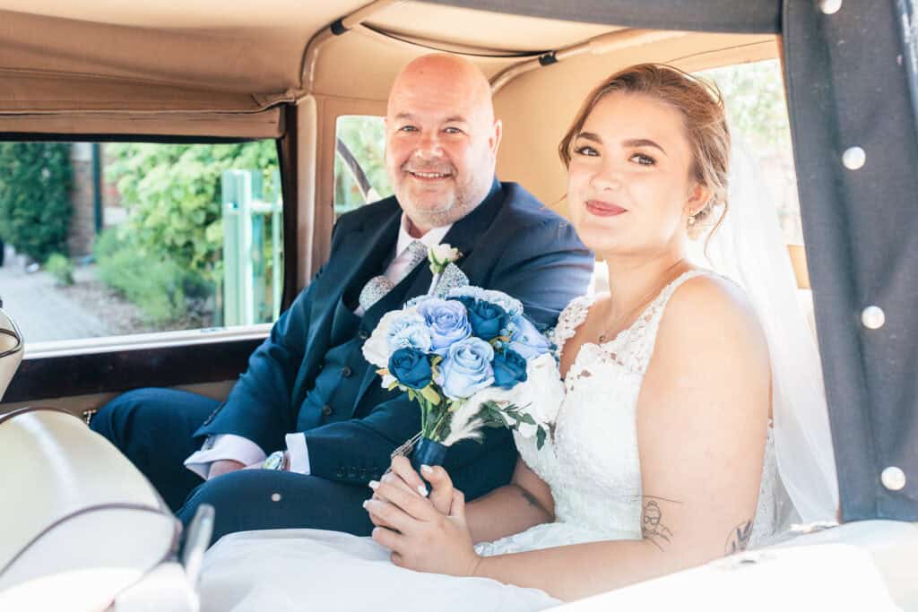 A bride in a white dress holding a bouquet of blue and white flowers sits beside a man in a suit, both smiling inside a vintage car. Sunlight brightens the scene through the open window.