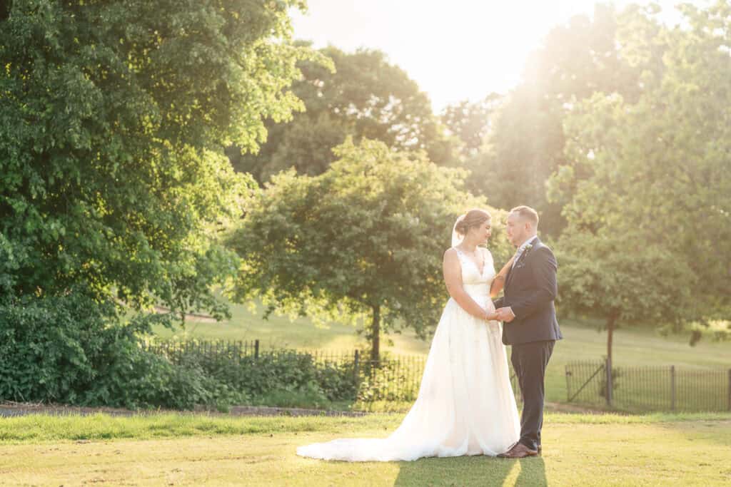 A bride and groom stand facing each other, holding hands on a sunny day in a green park, surrounded by trees and bathed in warm, golden sunlight.