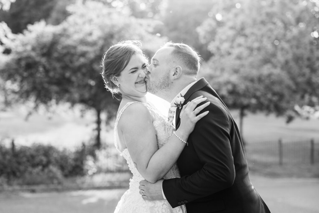 A joyful bride and groom share a playful moment outdoors, with the groom affectionately kissing the bride on the cheek. Both are smiling and dressed in wedding attire, surrounded by soft sunlight and blurred trees in the background.