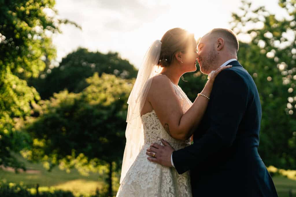 A bride and groom embrace and share a kiss outdoors at sunset, with sunlight glowing behind them. The bride wears a lace gown and veil, and the groom is in a dark suit. Trees and greenery surround them.