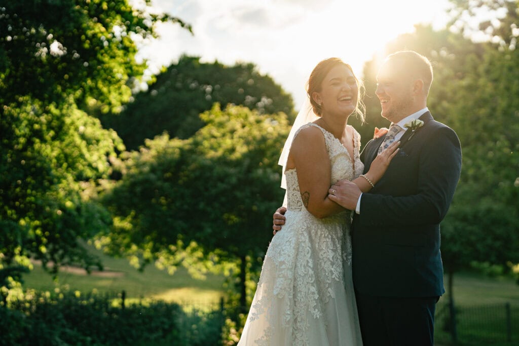 A bride and groom laugh together outdoors, bathed in warm sunlight, with lush green trees in the background. The bride wears a lace dress and veil, and the groom is in a dark suit and bow tie.