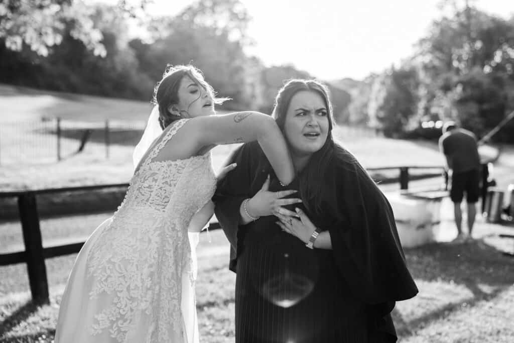 A bride in a lace wedding dress playfully pretends to punch a woman in a dark dress, who looks surprised. They are outdoors in sunlight, with trees, a fence, and a person in the background.