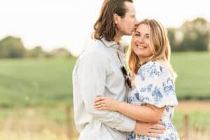 A man with long hair kisses a smiling woman on the forehead as they embrace in a sunlit field. The woman wears a blue and white floral dress; the background is green and softly blurred.