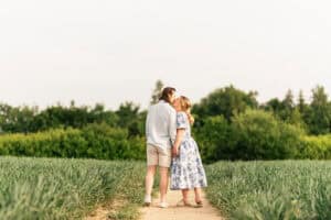 A couple stands on a dirt path in a green field, holding hands and sharing a kiss. The woman wears a blue and white floral dress, and the man wears a light shirt and shorts. Trees and greenery fill the background.