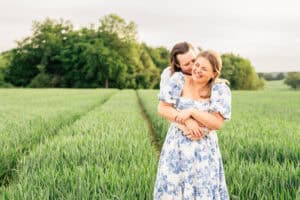 A happy couple stands in a green field. One person hugs the other from behind, both smiling and laughing. The sky is light and trees are in the background. The front person wears a white and blue floral dress.