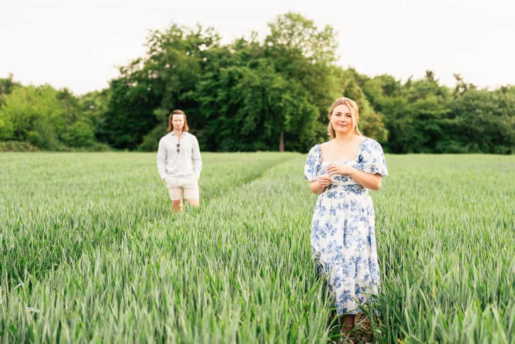 A woman in a blue and white floral dress stands smiling in a green field, while a man in a light shirt and shorts stands behind her on a path, with trees in the background.