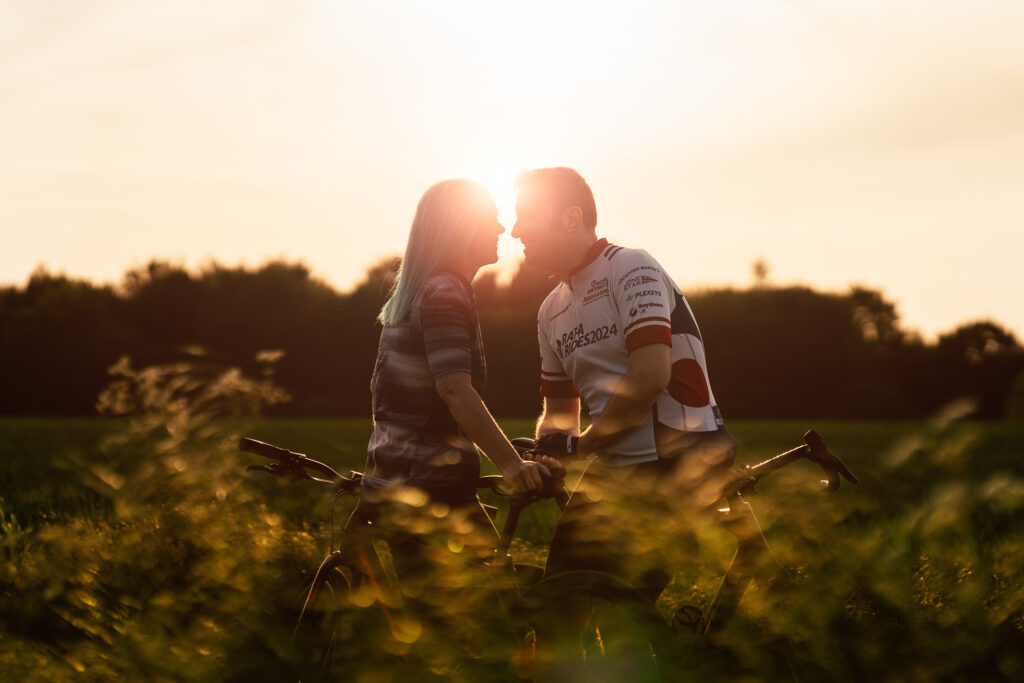 A couple stands with bicycles in a field at sunset, facing each other closely as if about to kiss, with sunlight glowing behind them and tall grass in the foreground.