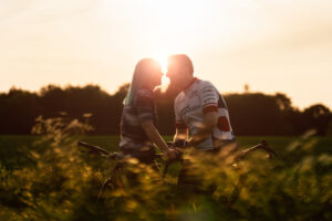 A couple stands with bicycles in a field at sunset, facing each other closely as if about to kiss, with sunlight glowing behind them and tall grass in the foreground.