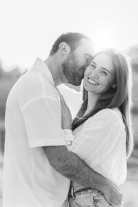 A man kisses a smiling woman on the cheek while they embrace outdoors. Both wear light-colored casual clothes, and sunlight softly highlights them from behind.