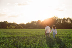 A couple holding hands walks through a green field at sunset, with trees in the background and sunlight streaming through the sky.