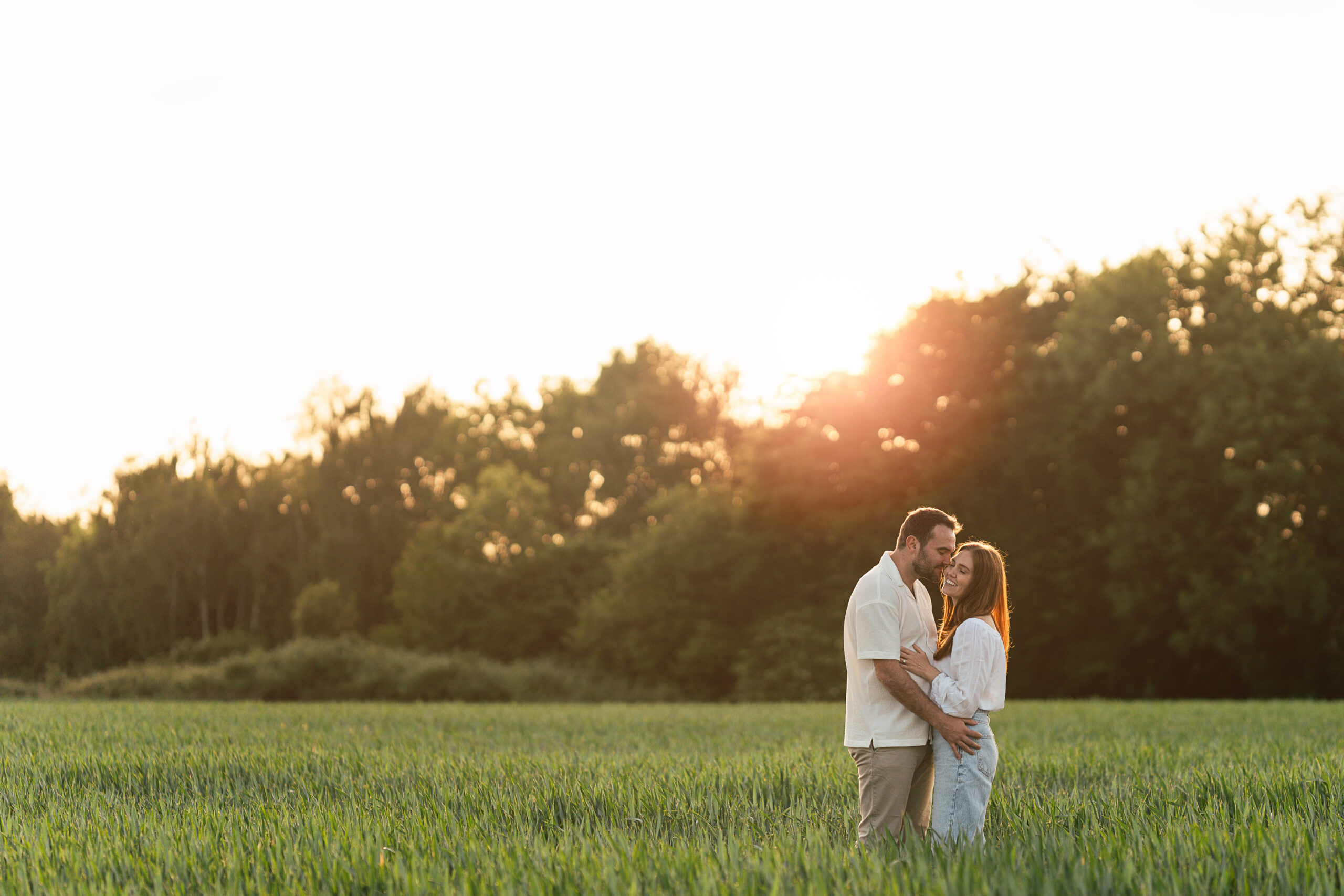 A couple stands closely together in a grassy field at sunset, embracing and smiling at each other, with trees and a bright sun in the background.