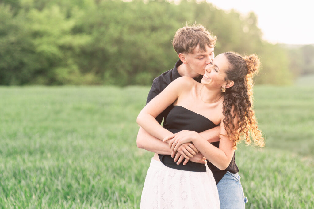 A young couple stands in a green field; the man hugs the woman from behind and playfully kisses her cheek. Both are smiling and appear happy, with soft sunlight and trees in the background.