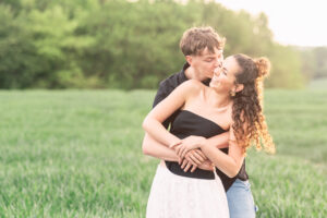 A young couple stands in a green field; the man hugs the woman from behind and playfully kisses her cheek. Both are smiling and appear happy, with soft sunlight and trees in the background.