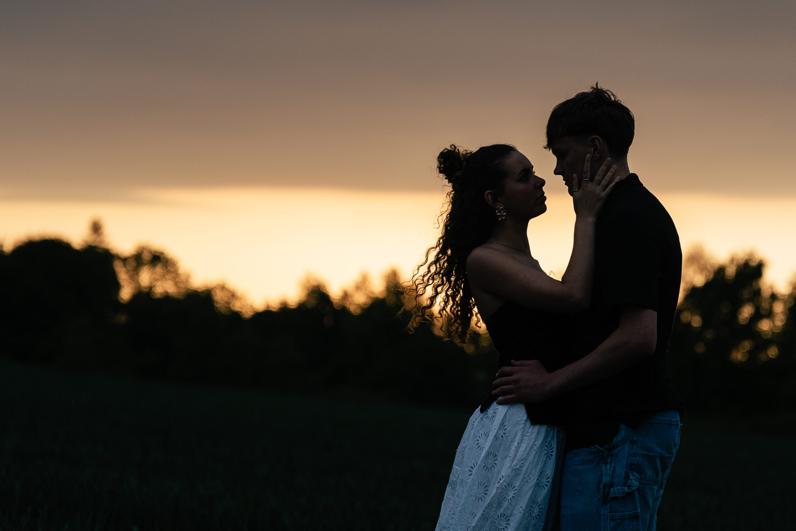 MJ020525-171 A couple stands close together in a field at sunset, facing each other. The woman gently holds the man's face as they gaze into each other's eyes, silhouetted against the golden sky and trees in the background.