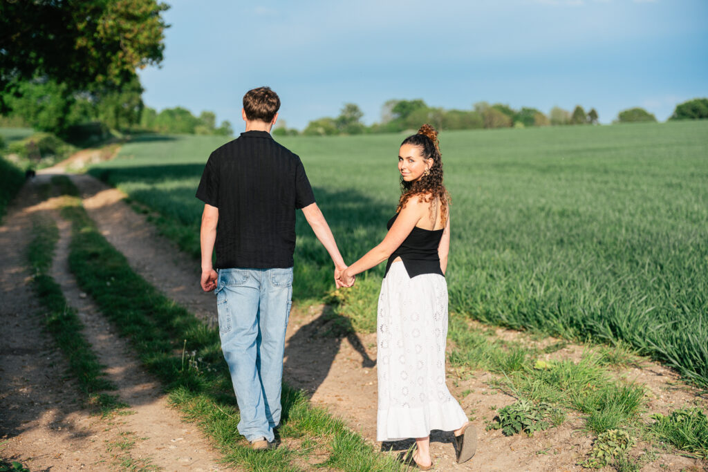 A couple walks hand in hand down a dirt path beside a green field; the woman looks back and smiles at the camera while the man faces forward.