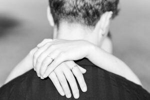 A close-up, black-and-white photo showing a person from behind with another person's arms wrapped around their neck. Both people wear rings on their fingers.
