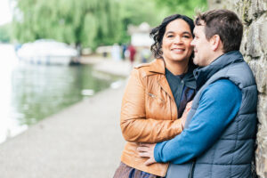 A smiling couple stands close together by a riverside path. The woman wears a brown leather jacket and the man wears a blue jacket. They lean against a stone wall with water and trees in the background.