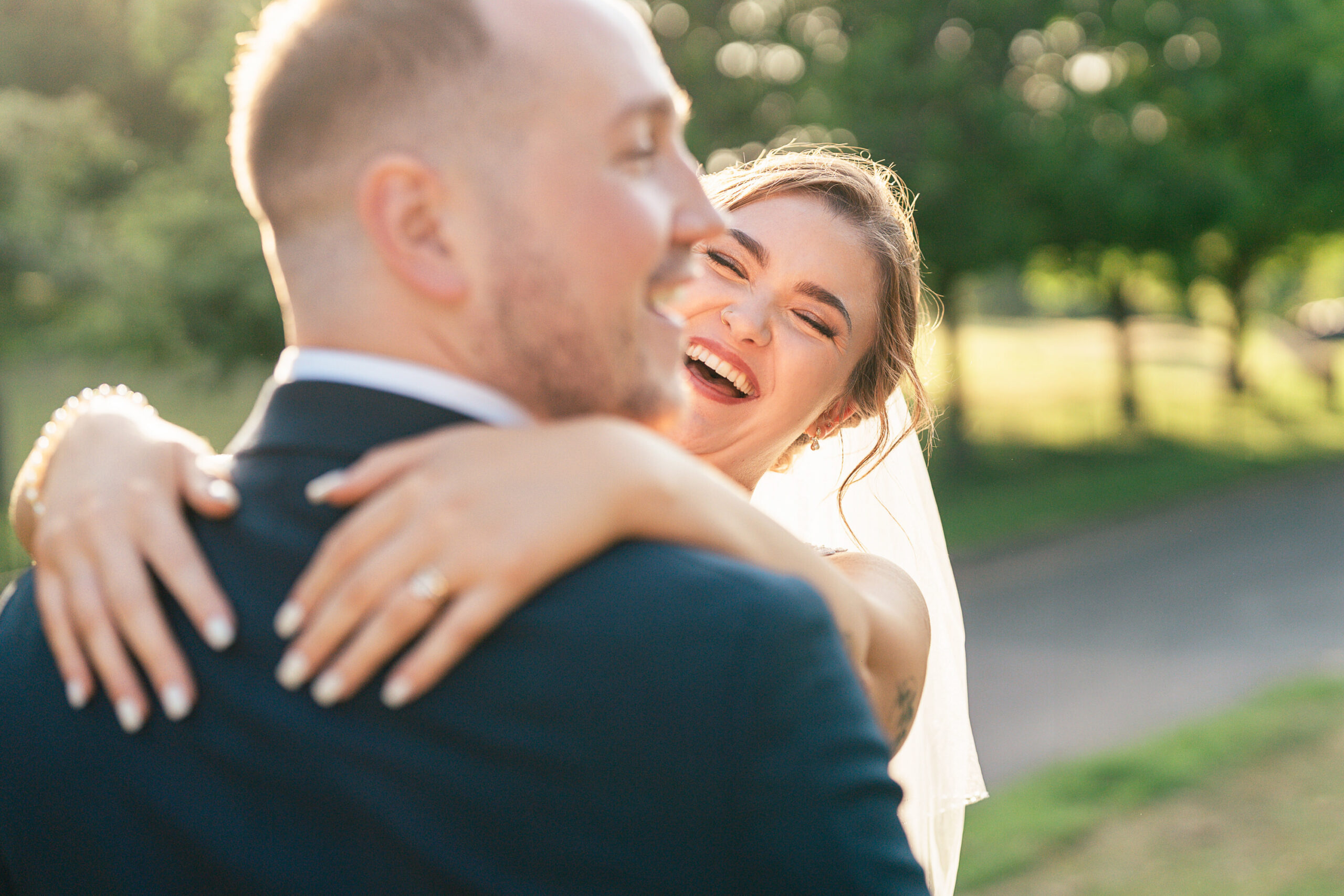 Over Shoulder A bride in a veil smiles joyfully with her arms around a groom, who is wearing a suit, as they embrace outdoors on a sunny day with greenery in the background.
