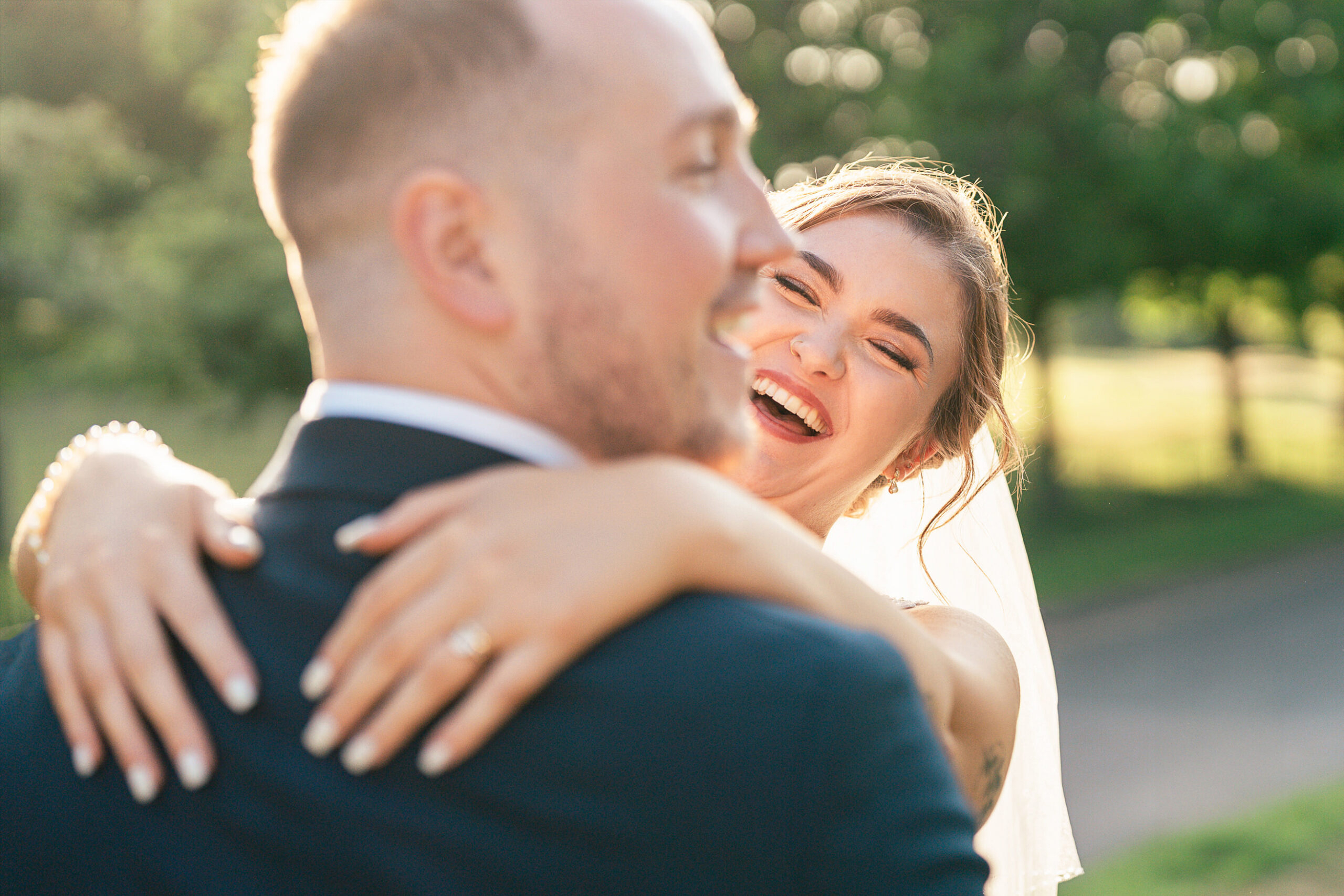 Over-ShoulderFINAL A bride with a veil smiles joyfully while hugging a groom outdoors, bathed in warm sunlight and surrounded by greenery.