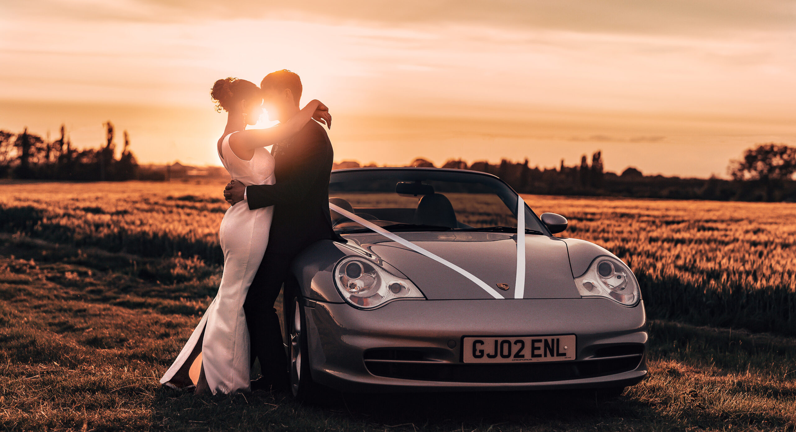 310525-129 A bride and groom embrace in front of a silver convertible decorated with white ribbons at sunset in a field, with soft golden light creating a romantic atmosphere.