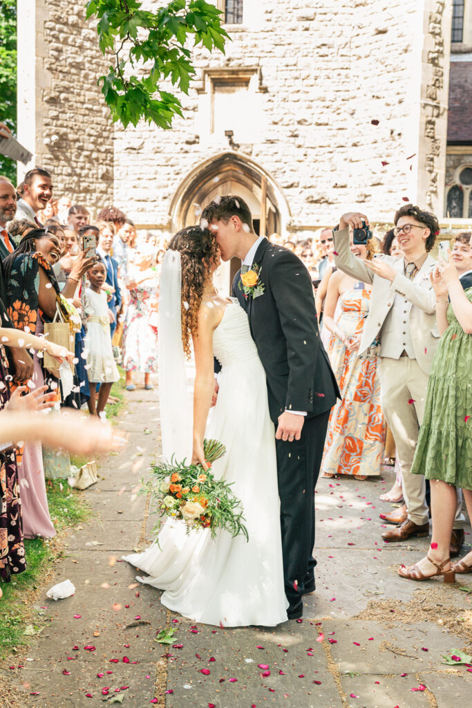 A bride and groom kiss outside a stone church as guests cheer, throw confetti, and take photos. The bride holds a bouquet and wears a white dress; the groom is in a black suit. The scene is joyful and sunlit.