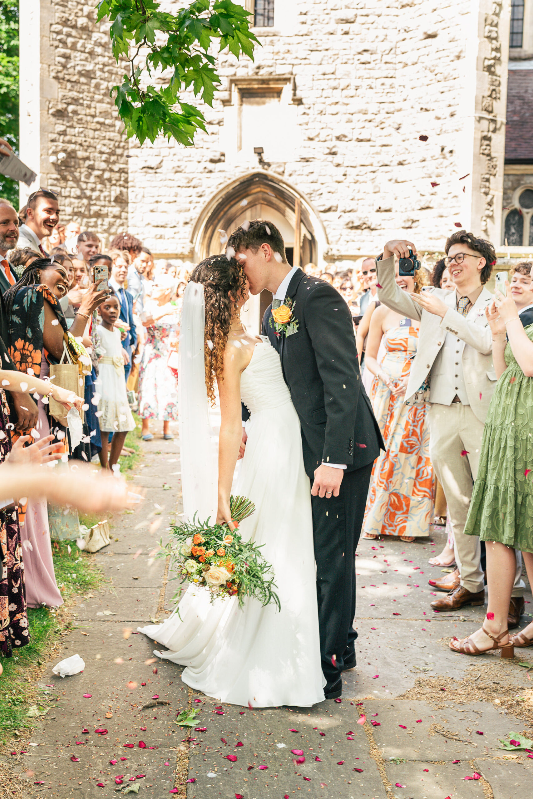 3835953777 A bride and groom kiss outside a stone church while wedding guests on both sides cheer, throw confetti, and take photos. The bride holds a bouquet and wears a long white dress; the groom is in a black suit.