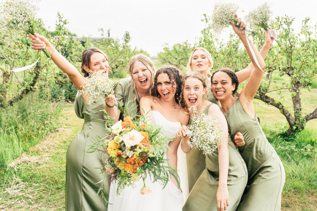 A joyful bride in a white dress stands with five bridesmaids in sage green dresses, all smiling and holding bouquets, celebrating outdoors in a lush, green orchard.
