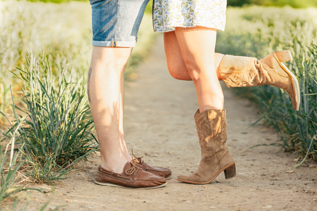 Close-up of a couple standing on a dirt path in a field. The woman wears a floral dress and cowboy boots, lifting one leg, while the man wears shorts and brown shoes. Their faces are not visible.