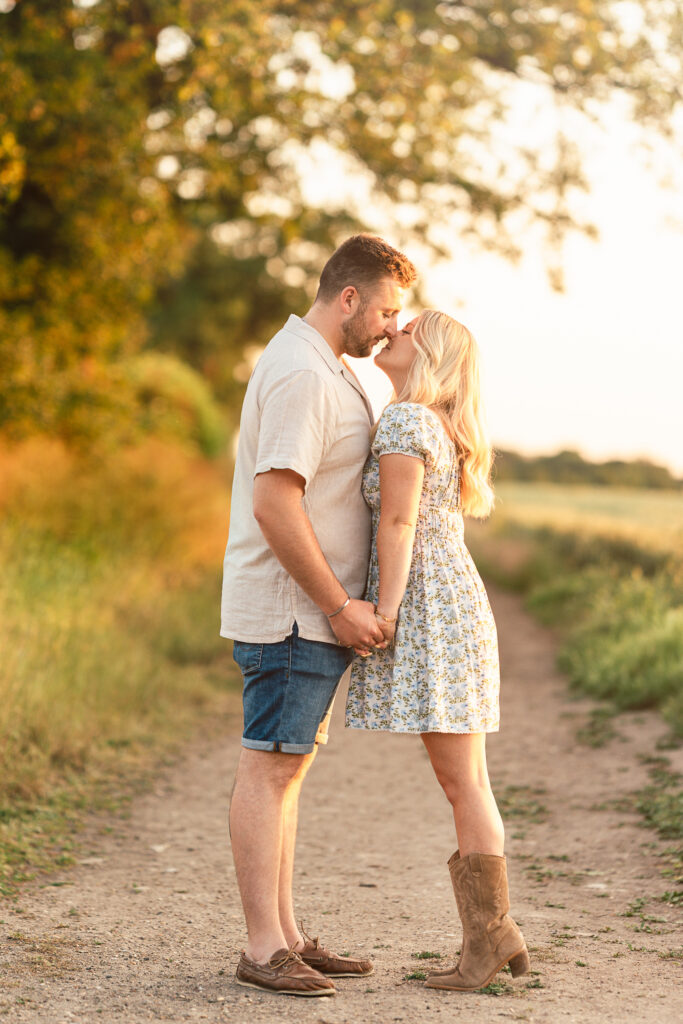 A couple stands on a dirt path, holding hands and facing each other. The man gently kisses the woman's forehead. They are outdoors with greenery and warm sunlight in the background.