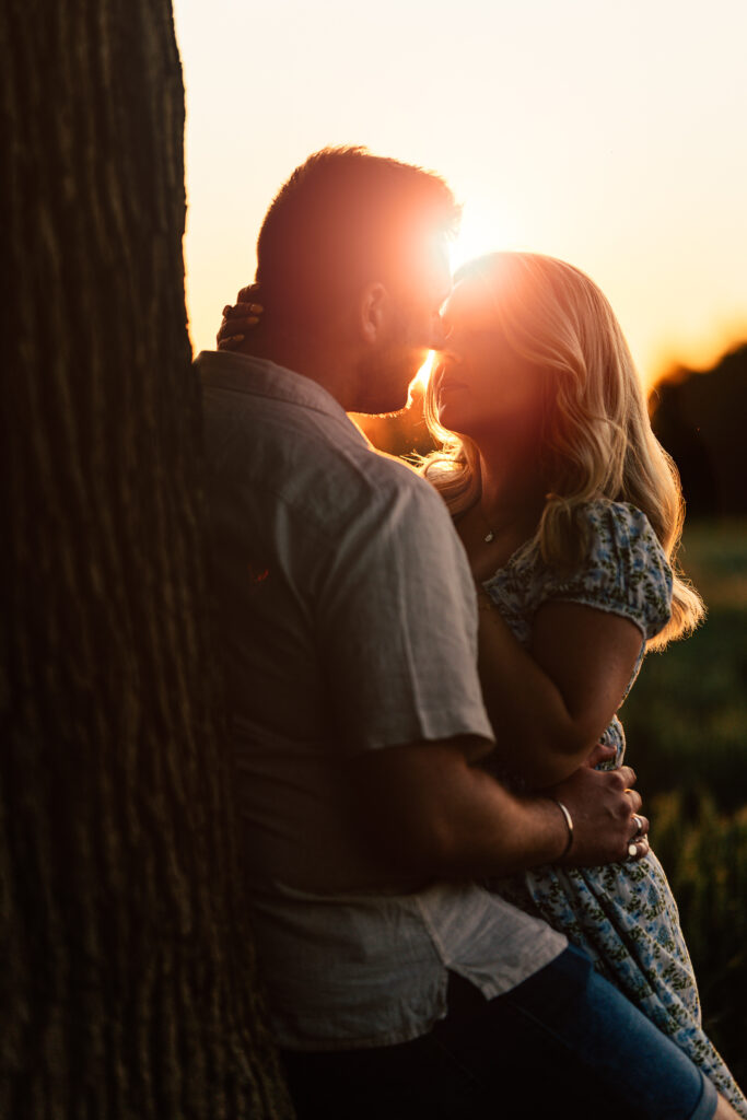 A couple embraces closely next to a tree at sunset, with golden sunlight shining between their faces, creating a warm, romantic atmosphere.