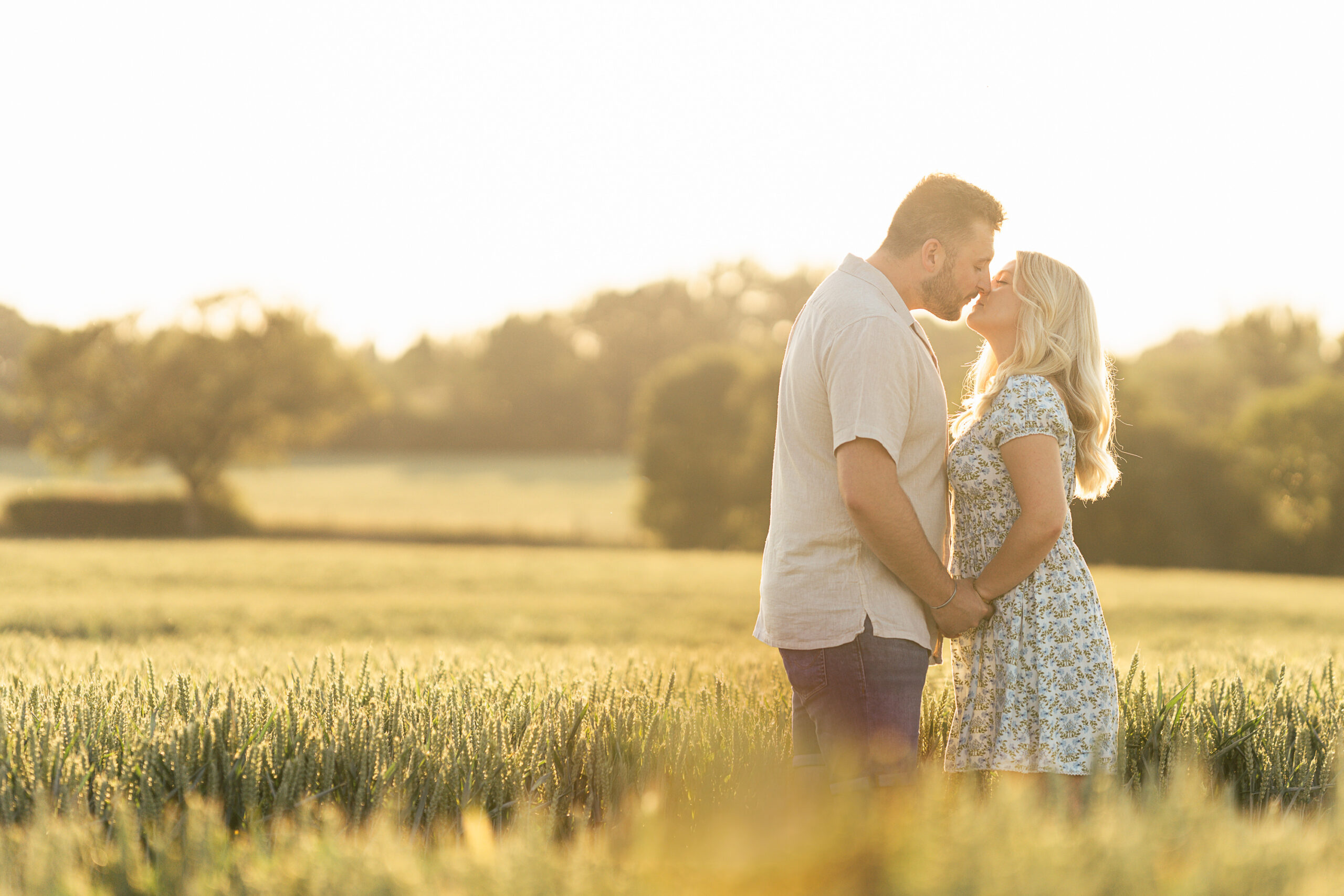 3852280732 A couple stands in a sunlit field, holding hands and facing each other. The man gently kisses the woman's forehead as they share an intimate moment surrounded by tall grass and a warm, golden glow.