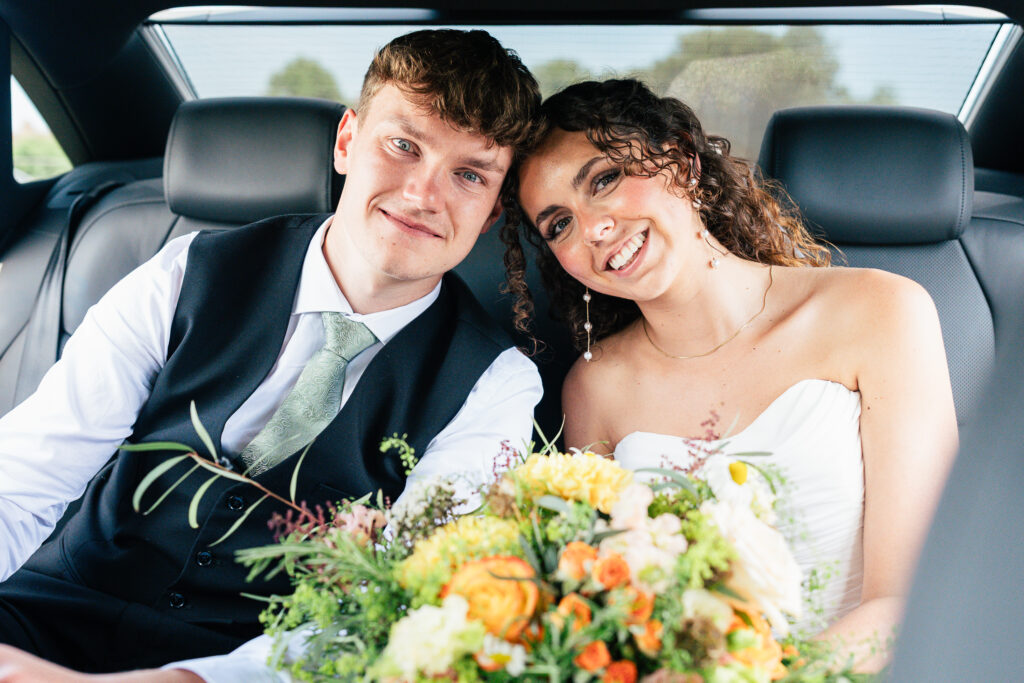 A smiling bride and groom sit closely together in the backseat of a car, holding a colorful bouquet of flowers. The bride wears a strapless white dress, and the groom is in a vest and tie.