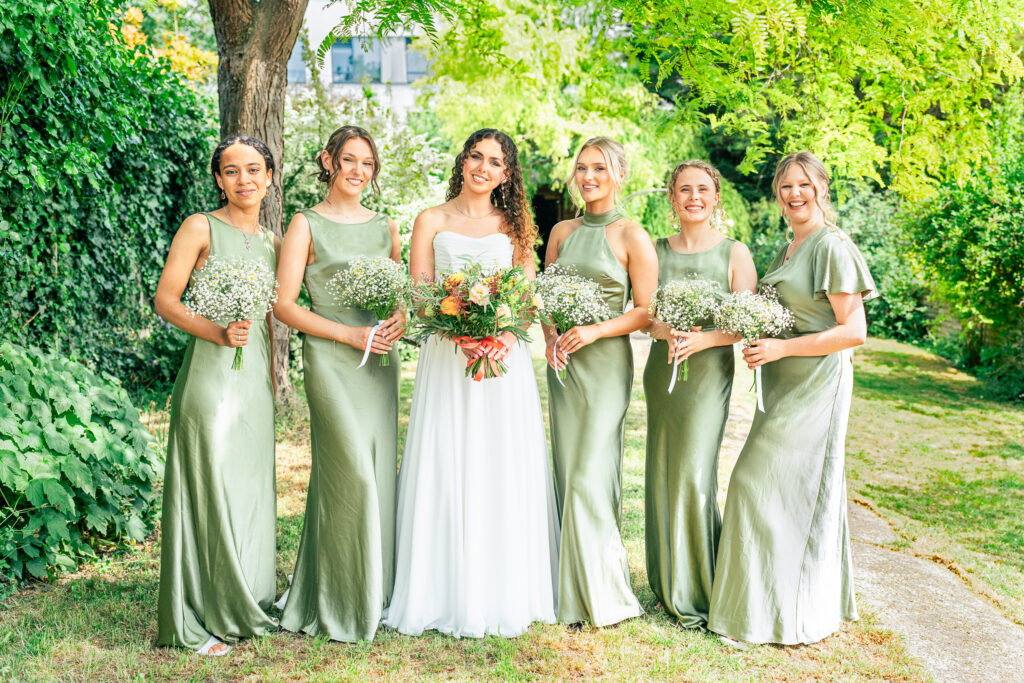 A bride in a white dress stands outdoors with five bridesmaids in matching sage green dresses, all holding bouquets and smiling in a lush, green garden setting.