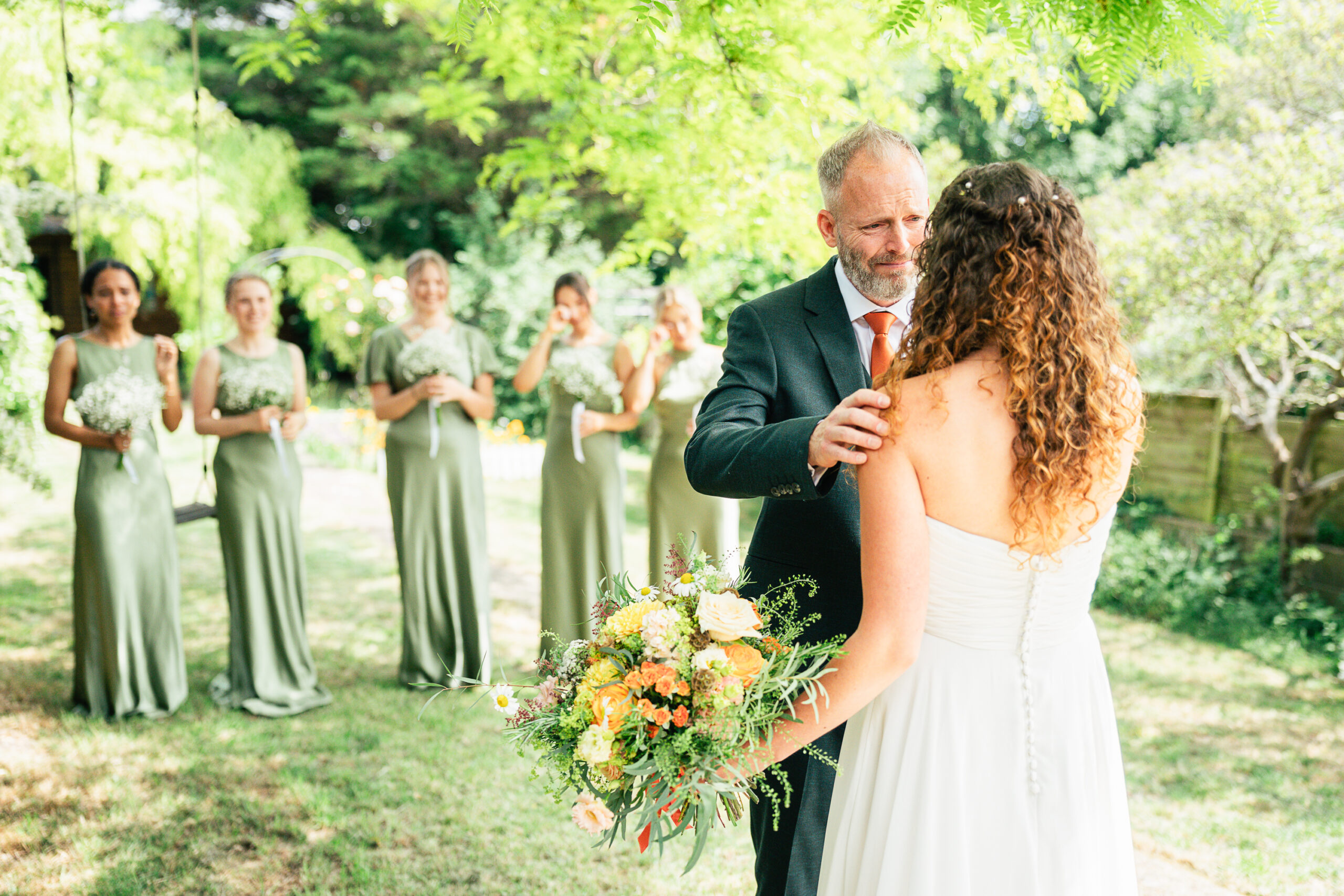 A man in a suit gently holds a bride’s shoulder during an outdoor wedding, with bridesmaids in sage green dresses smiling in the background. The scene is bright and surrounded by greenery.