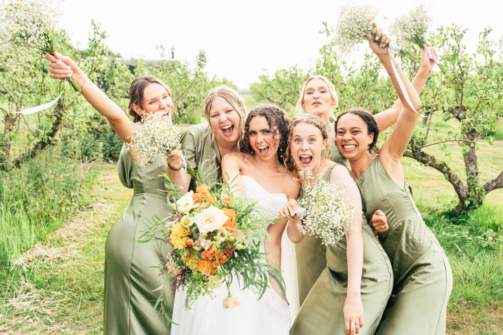 A joyful bride in a white dress stands outdoors with five bridesmaids in sage green dresses, all smiling and holding bouquets, celebrating together in a lush, green garden setting.