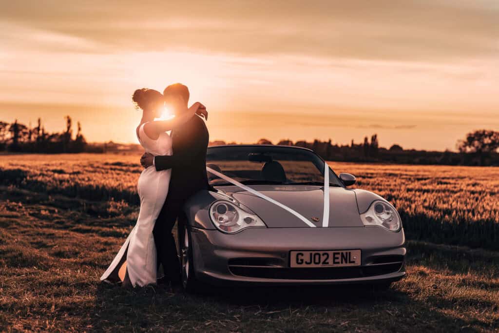 A couple in wedding attire embraces near a silver convertible decorated with white ribbons, parked in a field at sunset. The sun glows behind them, casting a romantic light on the scene.