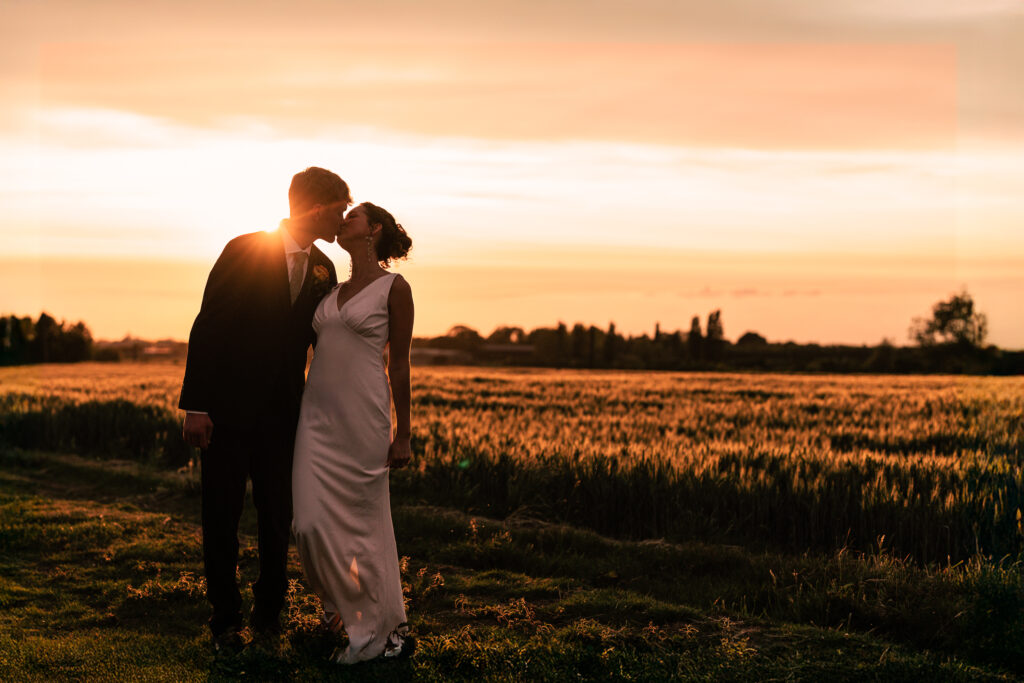 A couple in wedding attire stands in a field at sunset, sharing a kiss. The sun is low on the horizon, casting a warm golden glow over the landscape and creating a beautiful silhouette of the pair.