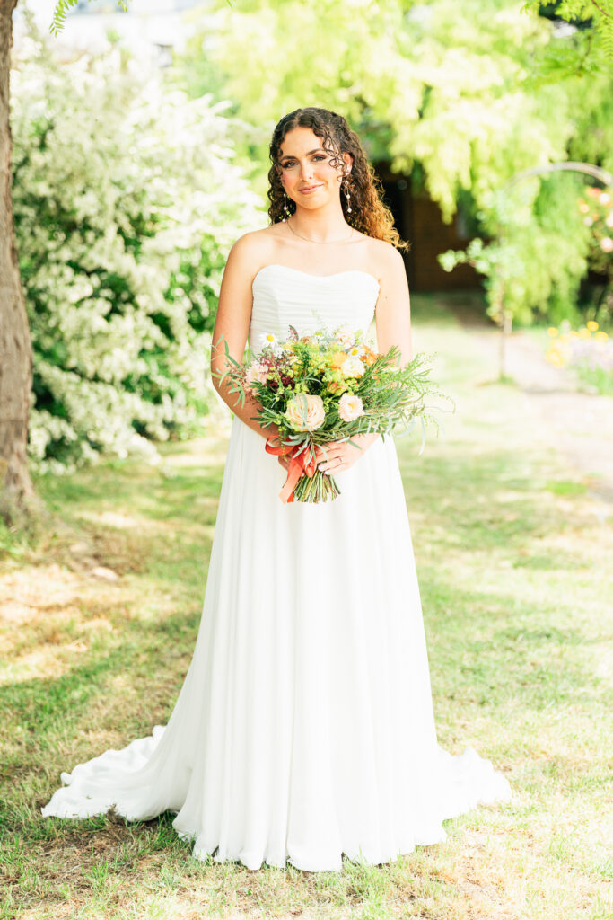 A woman in a strapless white wedding dress stands outdoors on grass, holding a bouquet of flowers. Lush green trees and bushes are in the background, and sunlight filters through the leaves.