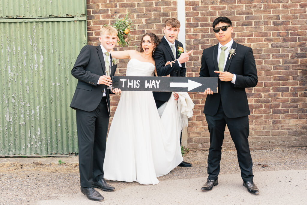 A bride and three groomsmen in suits pose playfully outside by a brick wall, holding a black sign with a white arrow and the words “THIS WAY.” All are smiling and having fun.