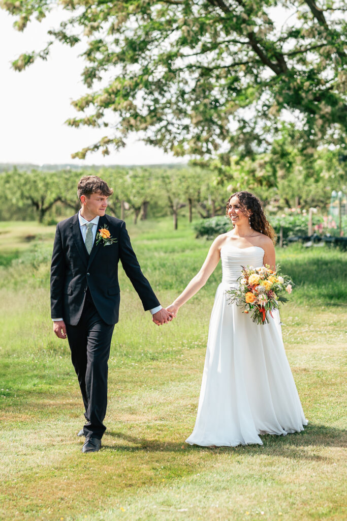 A bride in a white dress holding a colorful bouquet and a groom in a black suit hold hands and walk across a grassy field, surrounded by trees and greenery on a sunny day.