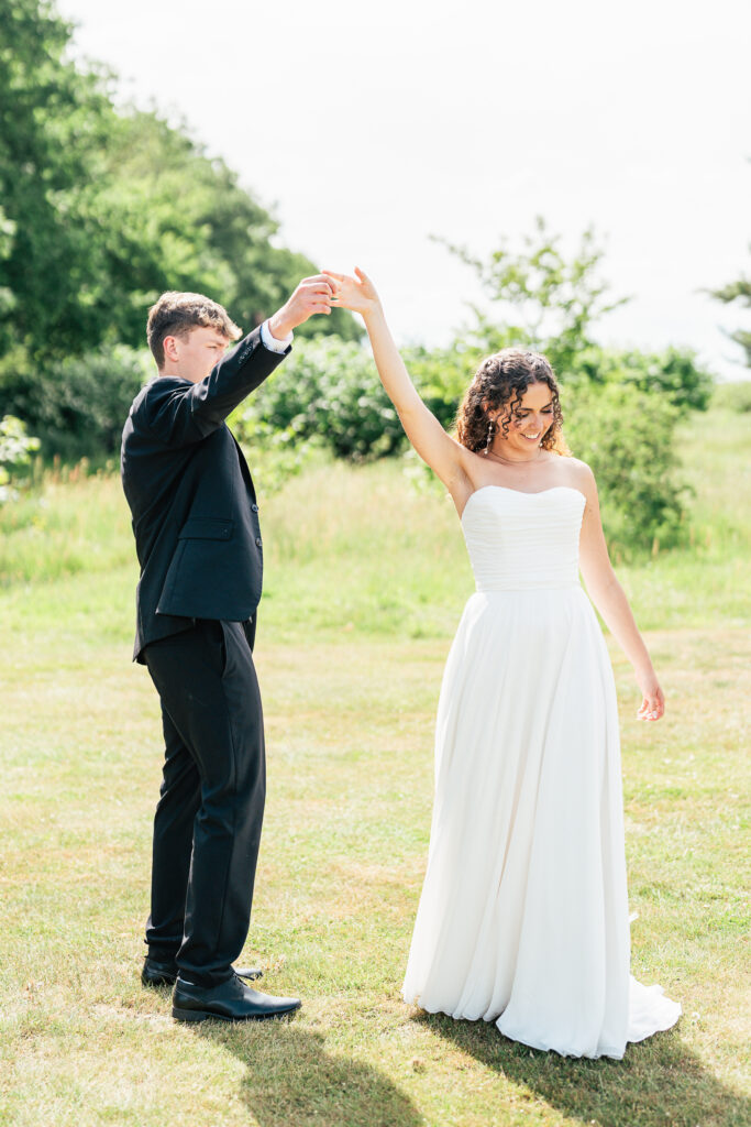 A bride in a white dress and a groom in a black suit dance together outdoors on a sunny day, holding hands and smiling, with green grass and trees in the background.