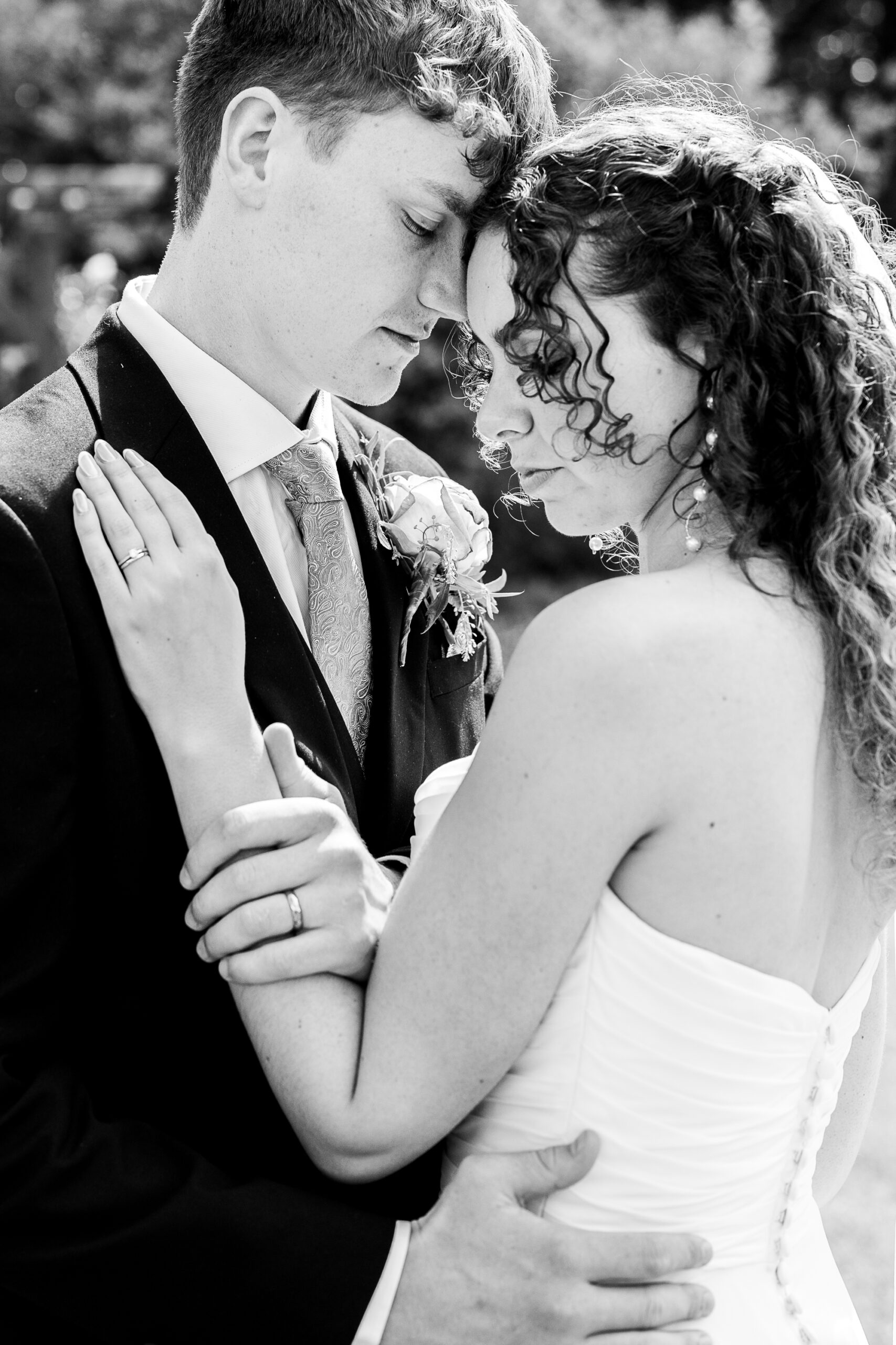 MJ310525-824 A bride and groom embrace closely outdoors, eyes closed and foreheads touching, sharing an intimate moment. The bride wears a strapless dress and the groom is in a suit with a boutonniere. Black and white photo.