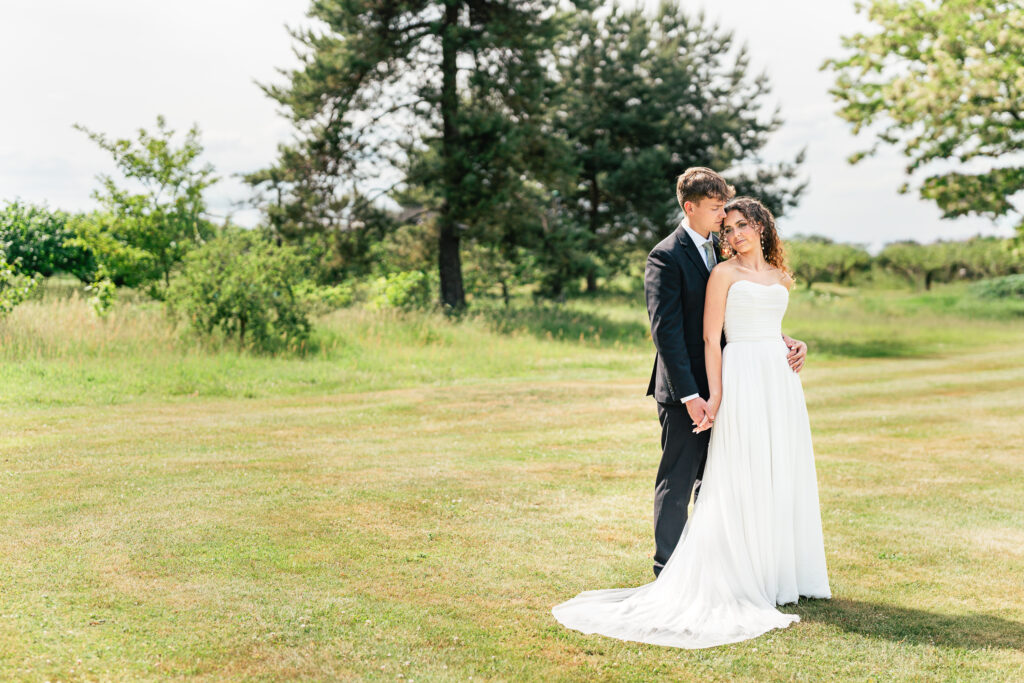 A bride in a white dress and a groom in a black suit stand closely together on a grassy field, surrounded by trees and sunlight, sharing an affectionate moment.