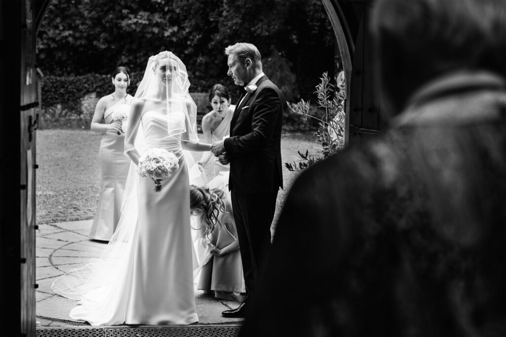 A bride in a veil and gown stands holding a bouquet beside a man in a tuxedo at a church entrance, with bridesmaids adjusting her dress in the background. The photo is in black and white.