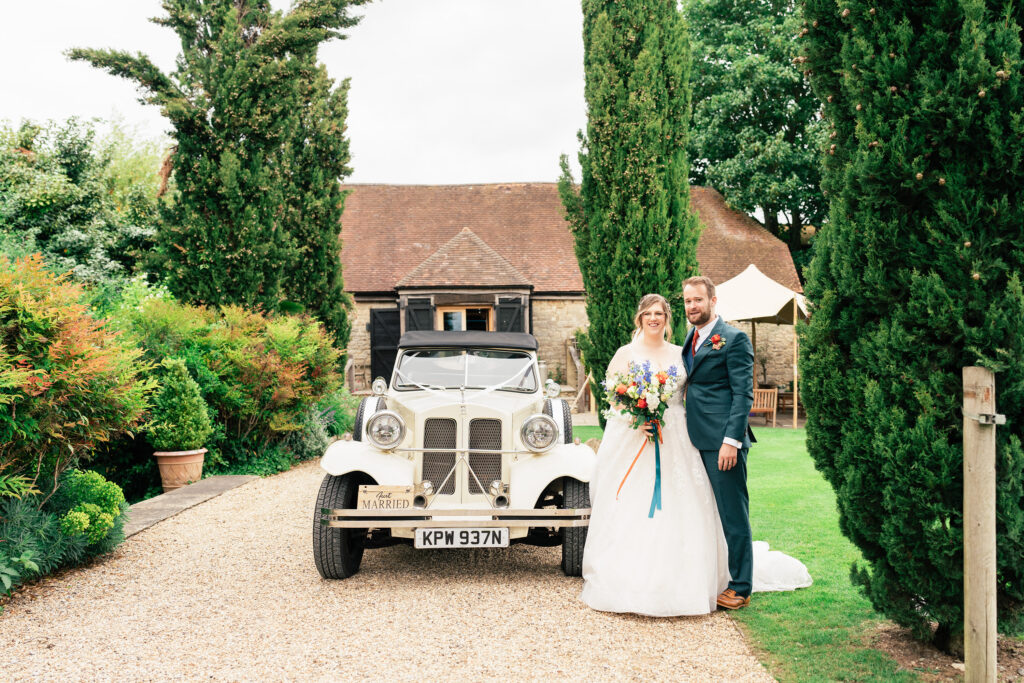 A bride and groom stand together in front of a vintage white car with a “Married” sign, outside a rustic building surrounded by greenery and tall trees.