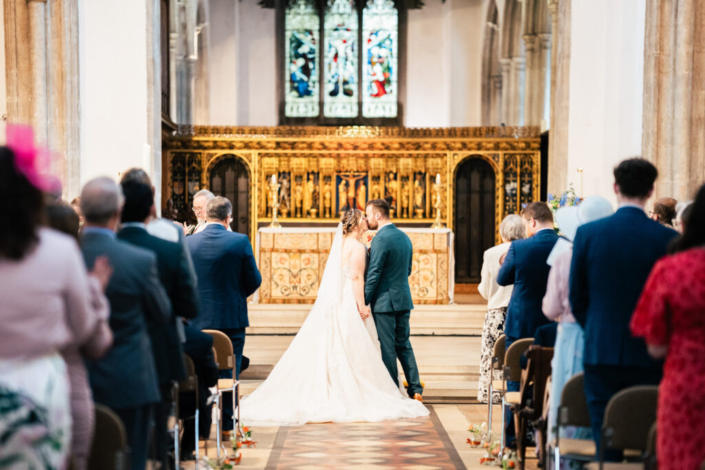 A bride and groom stand at the altar in a church, holding hands and facing stained glass windows, while wedding guests sit and watch from both sides of the aisle.