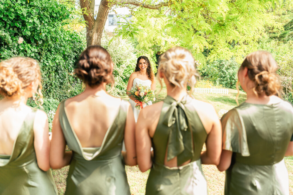A bride in a white dress holding a bouquet stands outdoors, smiling at four bridesmaids in matching green dresses who are facing her with their backs to the camera. Lush greenery and sunlight surround them.
