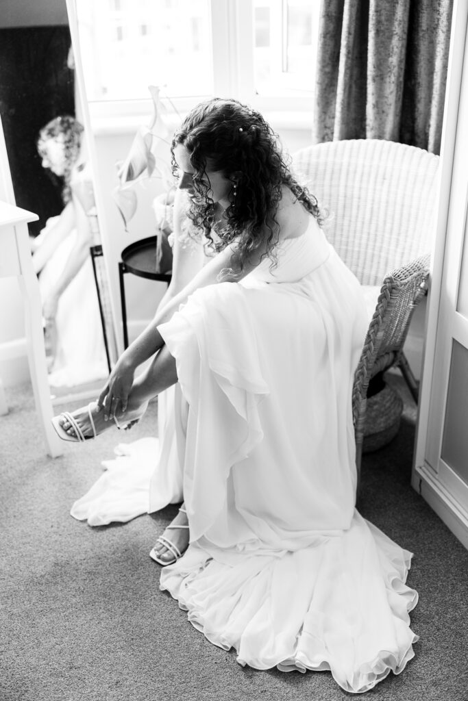A bride in a flowing wedding dress sits on a wicker chair, bending forward to adjust her sandal. Her reflection is visible in a nearby mirror. The scene is softly lit, creating an intimate, serene atmosphere.