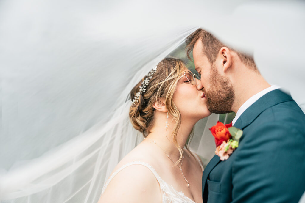 A bride and groom share a kiss under a sheer white veil. The bride wears glasses, a jeweled hairpiece, and a white dress, while the groom wears a dark suit with a red boutonniere.